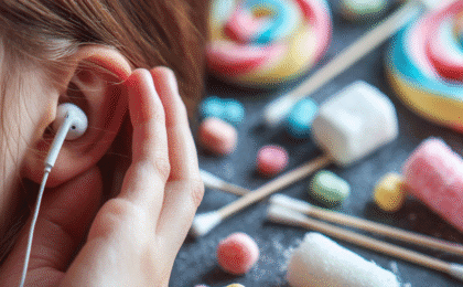Close-up of a person holding their ear while wearing white earphones with colorful candies in the background.